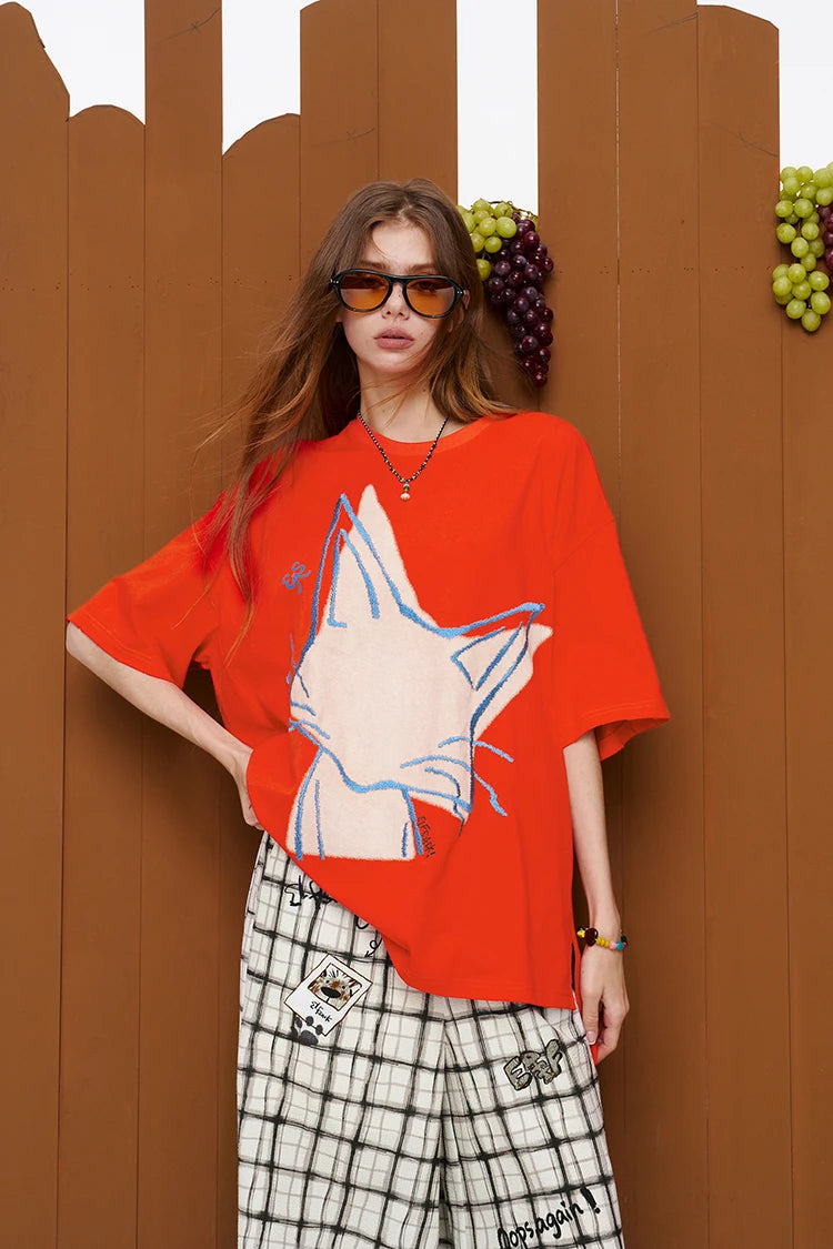 A woman wearing an oversized bright red graphic T-shirt with a large white fox design outlined in blue, paired with black-and-white plaid pants, standing against a brown wooden wall with decorative grape props.