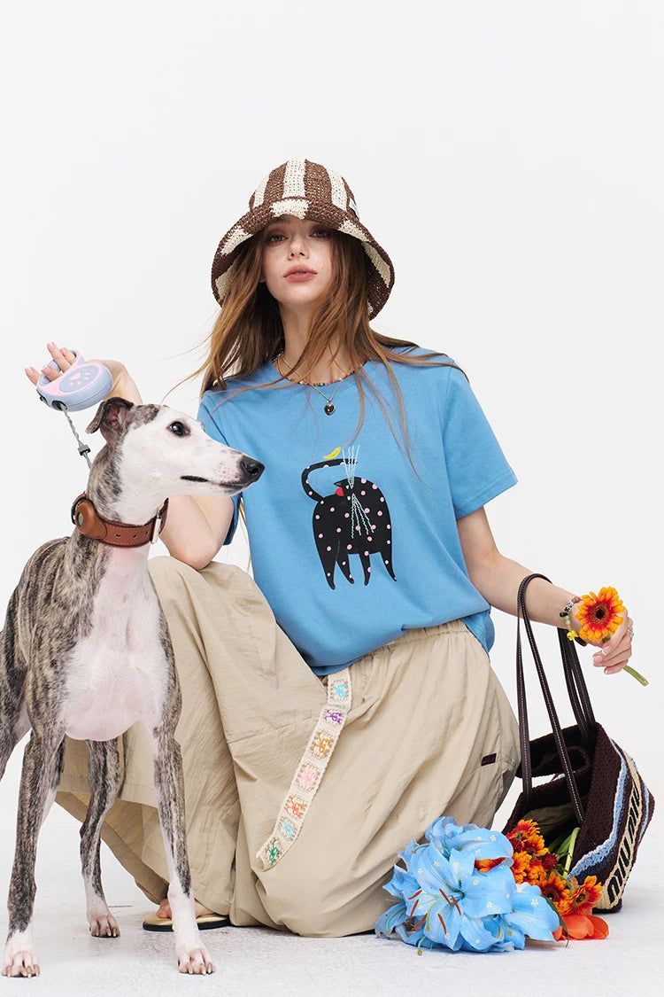 A woman wearing a blue graphic t-shirt, beige cargo pants, and a brown and beige bucket hat kneels next to a greyhound dog on a white studio background, holding a flower and a bag.