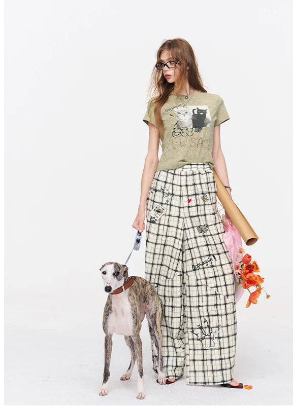 A woman modeling a graphic t-shirt and a black-and-white checkered wide-leg skirt, standing next to a greyhound dog on a white studio background. She holds a pink bag and orange flowers.