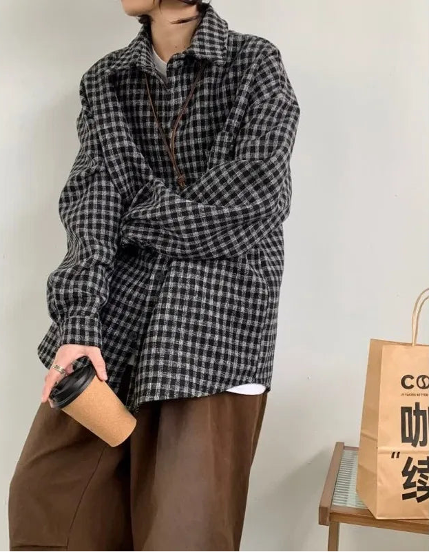 Person wearing a black and white checkered shirt with brown pants, holding a coffee cup, standing next to a wooden table with a bag on it.