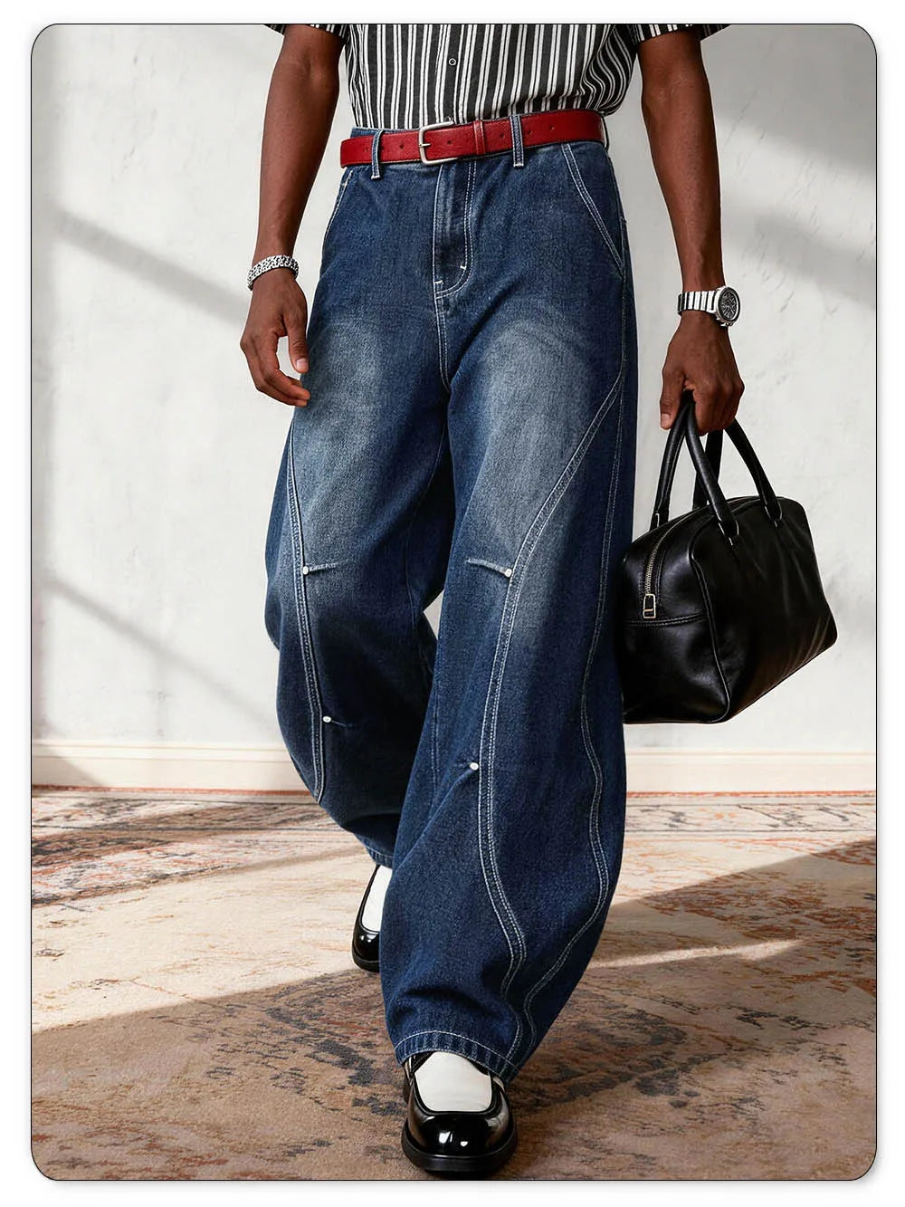 A person wearing oversized dark blue denim jeans with diagonal seam details, styled with a red belt, a black-and-white striped shirt, black shoes, and carrying a black handbag.