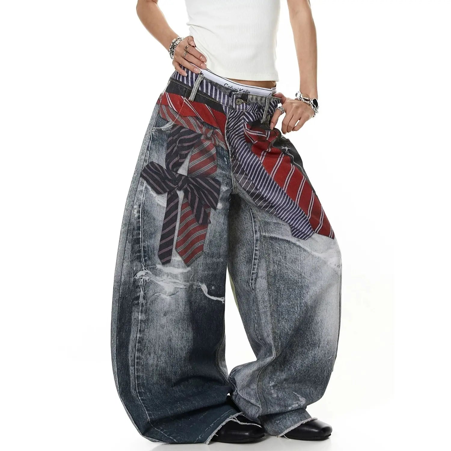 A person wearing extremely baggy distressed denim jeans with a printed pattern of oversized neckties in red, navy, and white, styled with a white tank top and black shoes against a white background.