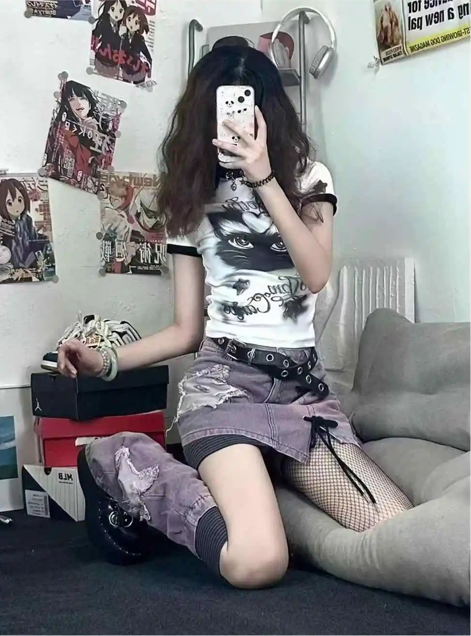 A person takes a mirror selfie in a room, wearing a white graphic t-shirt with a black cat print, a distressed purple denim mini skirt with a black lace-up belt, and fishnet stockings.