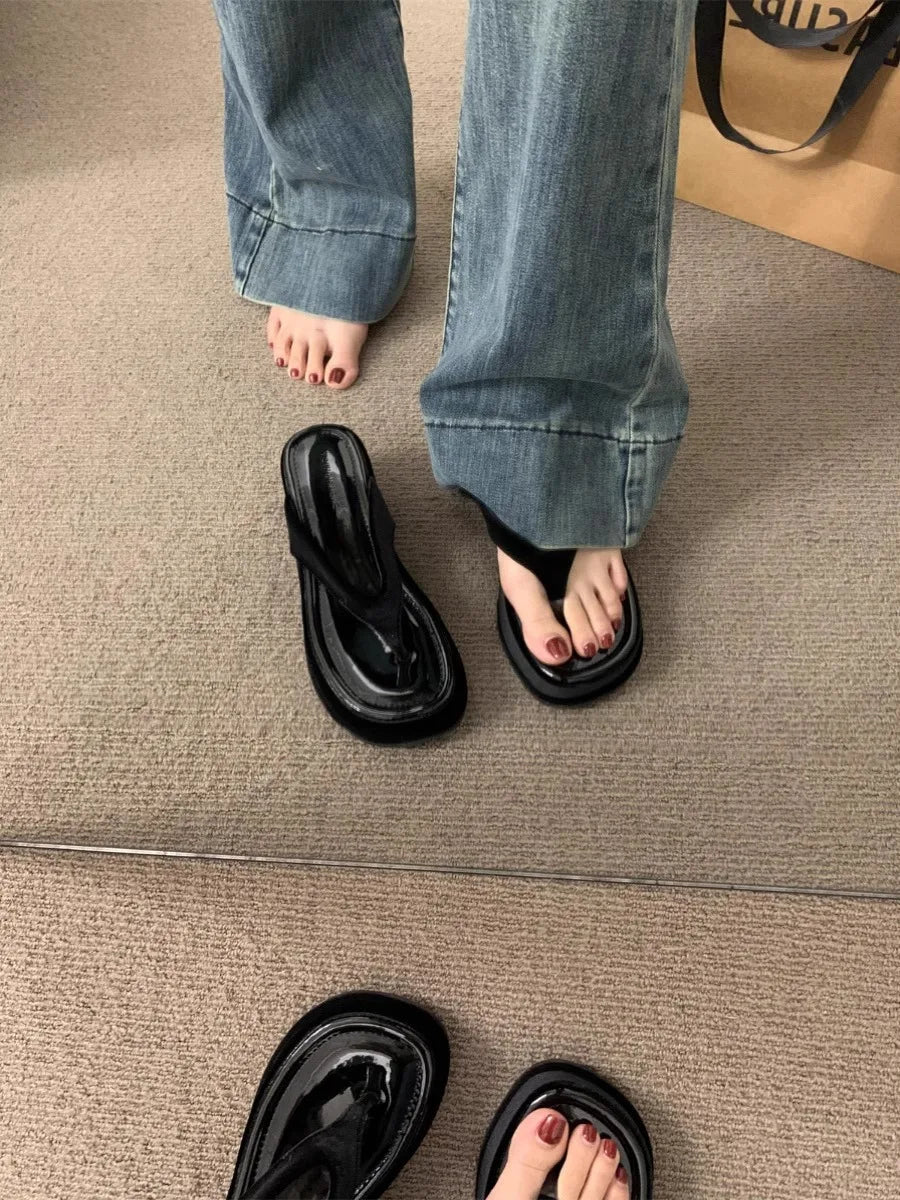 Black patent leather thong sandals with chunky platform soles, shown on a model's feet with blue jeans, with a mirror reflection showing additional pairs on a beige carpeted floor.