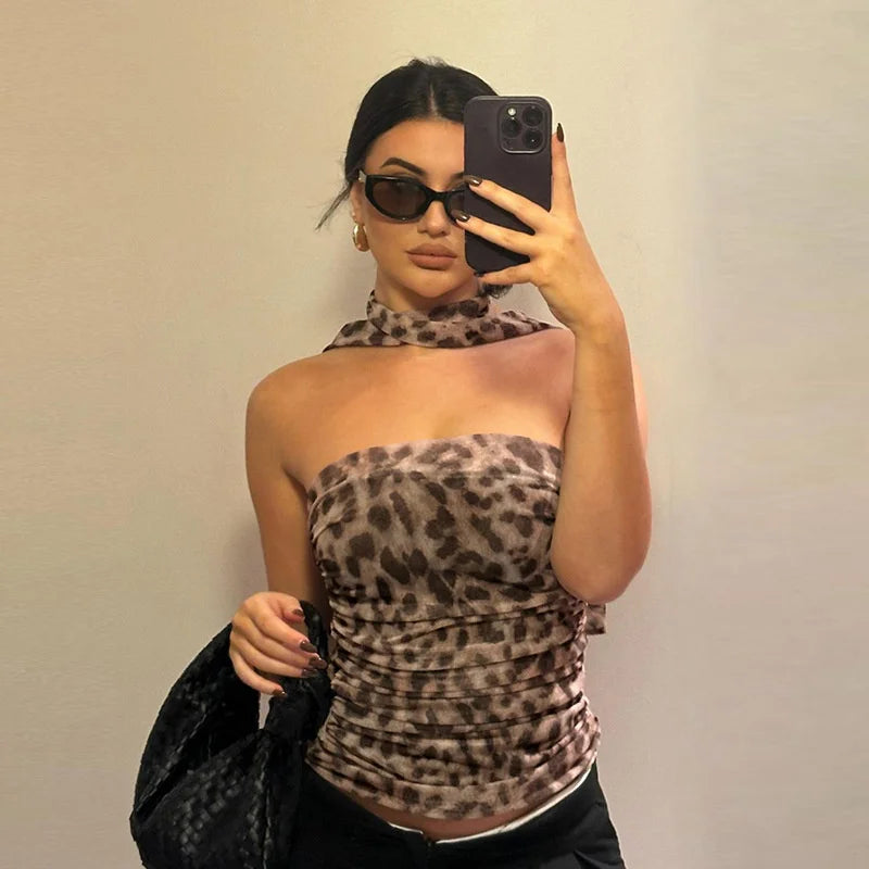 A woman takes a mirror selfie wearing a ruched leopard-print halter top, black sunglasses, and holding a black patterned handbag against a plain beige wall.