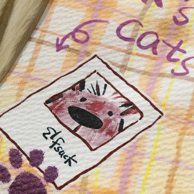 A close-up of a printed textile featuring a hand-drawn cat face inside a Polaroid-style frame, with purple paw prints and the word 'cats' on a plaid pattern background.