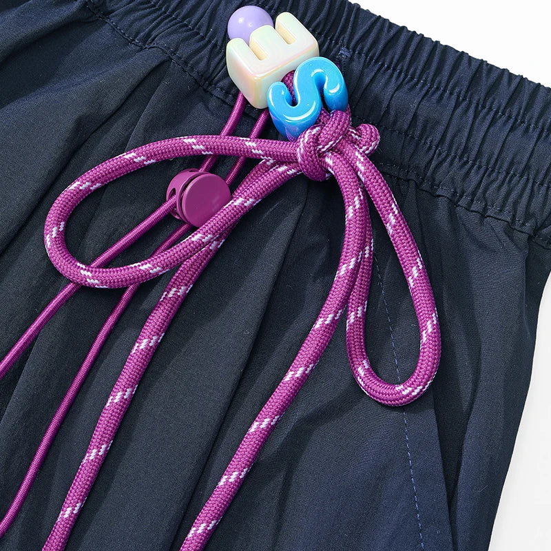 Close-up of a dark navy or black fabric garment with a purple drawstring and colorful plastic toggle hardware, including a blue S-shaped piece and a cream-colored piece.