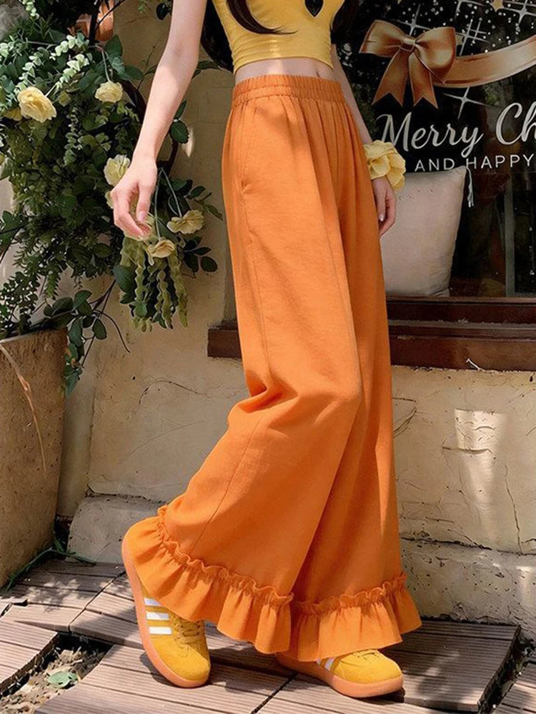 A person wearing bright orange wide-leg ruffled pants and yellow shoes, standing outdoors near a rustic wall and potted plants.