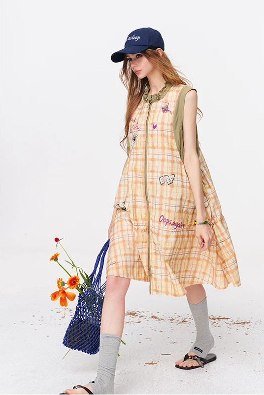 A woman models a sleeveless yellow and orange plaid dress with colorful embroidered patches, styled with a navy blue baseball cap, a blue mesh bag, gray socks, and sandals against a white studio background.
