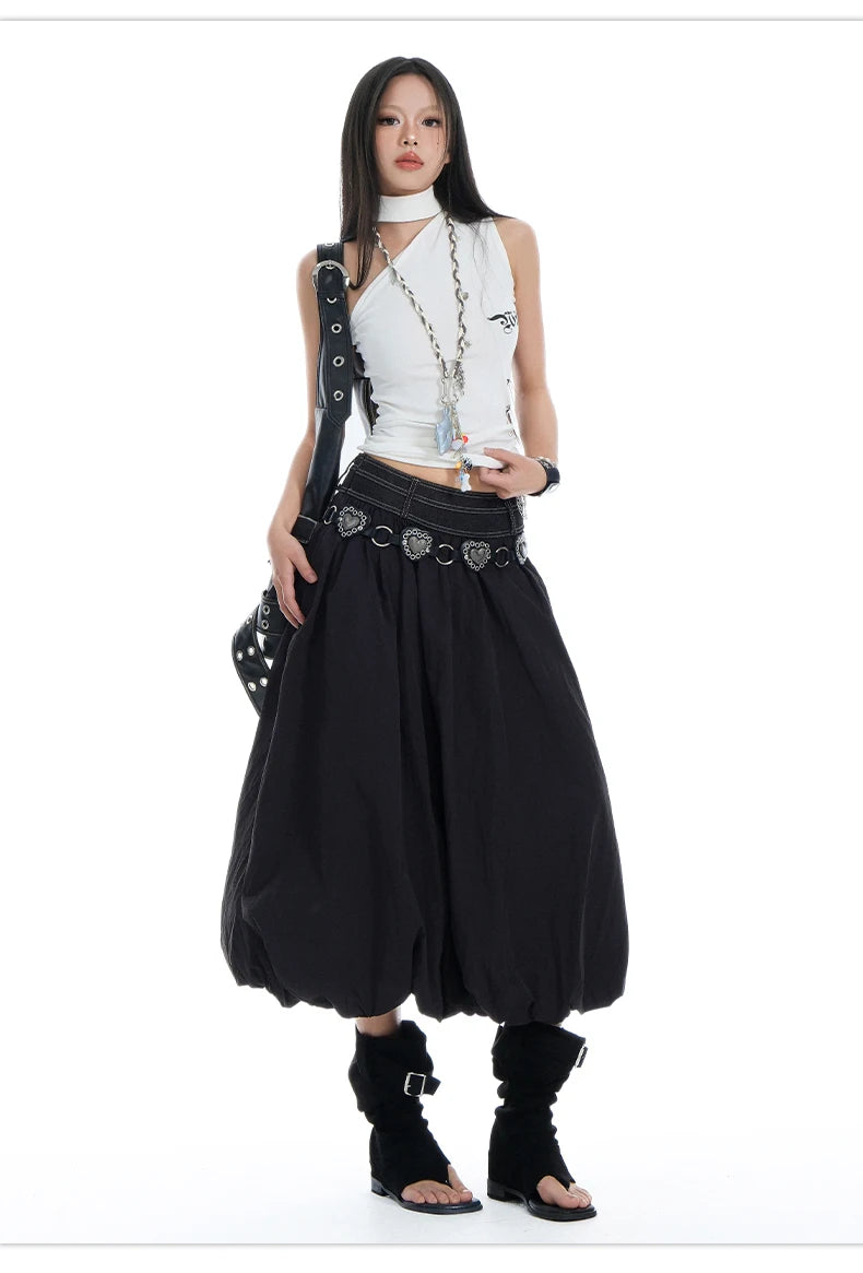 A model wearing a white sleeveless crop top, a long black skirt with a silver heart belt, black ankle boots, and carrying a black studded shoulder bag against a plain white background.
