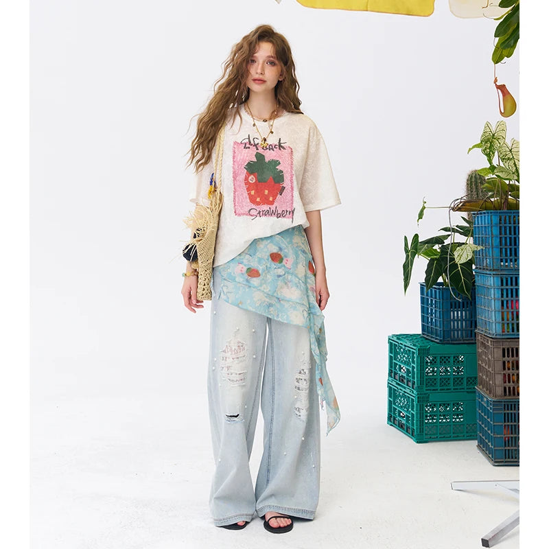 A woman stands in a studio wearing a cream graphic T-shirt with a strawberry print, light blue distressed wide-leg pants, and a sheer patterned wrap, with plants and stacked crates to the right.
