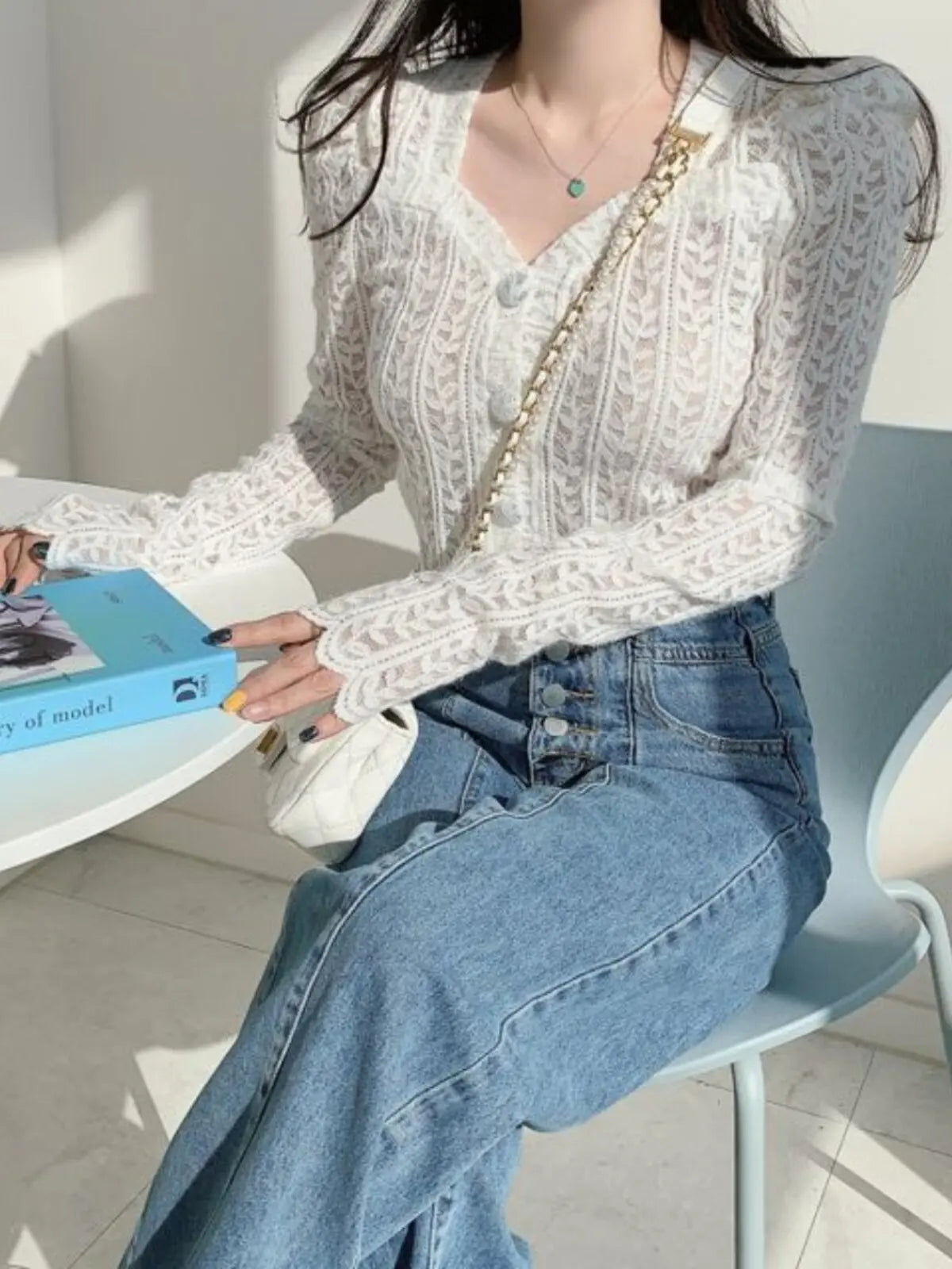 A woman seated indoors wearing a white lace long-sleeve cardigan with a V-neck and blue jeans, holding a light blue book and carrying a small white shoulder bag with a gold chain strap.