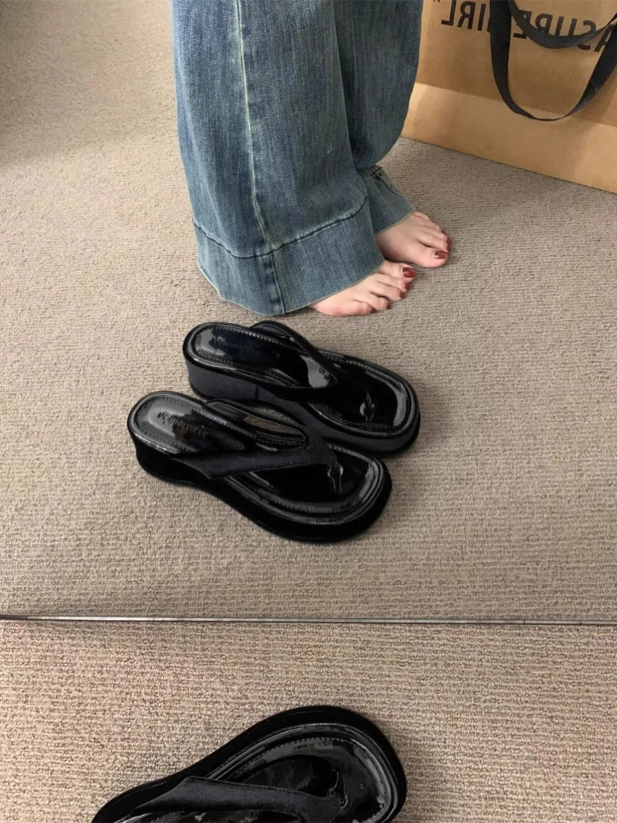 Black patent leather thong sandals with chunky platform soles, shown on a beige carpet next to a person's feet wearing blue jeans.