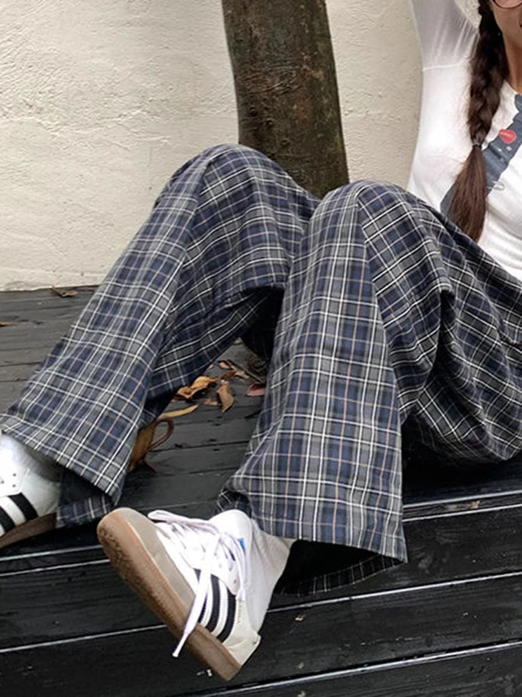 A person sitting on a dark wooden bench wearing wide-leg navy and gray plaid pants, white socks, white sneakers, and a white top.