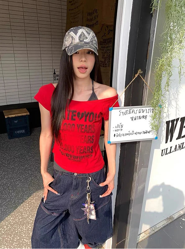 A woman wearing a red off-the-shoulder graphic t-shirt, dark blue baggy jeans, and a gray patterned baseball cap, standing in front of a storefront.