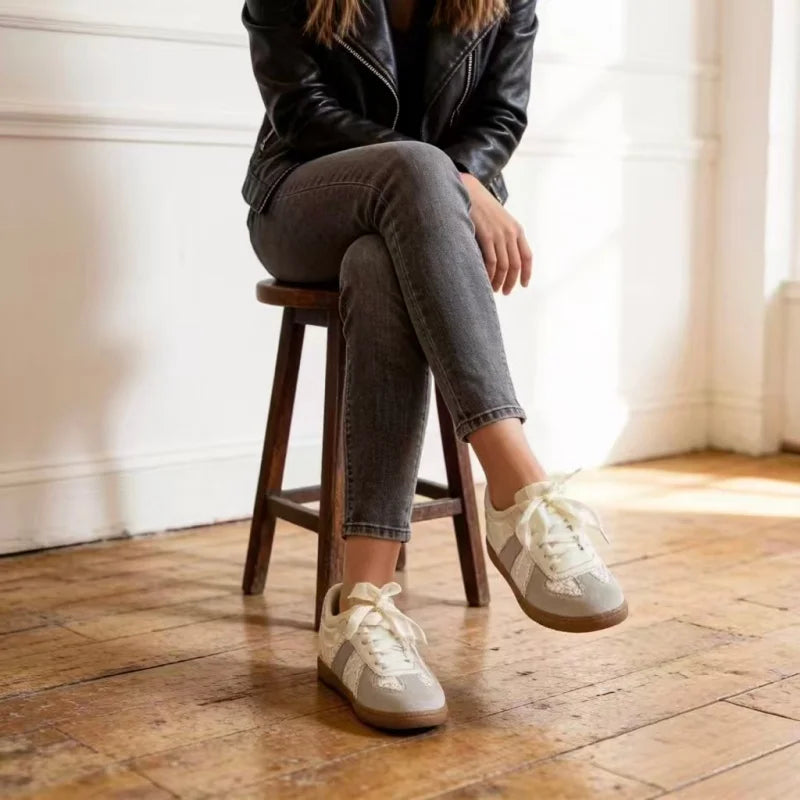 A person sitting on a wooden stool in a bright room, wearing gray skinny jeans, a black leather jacket, and light beige low-top sneakers with white laces.