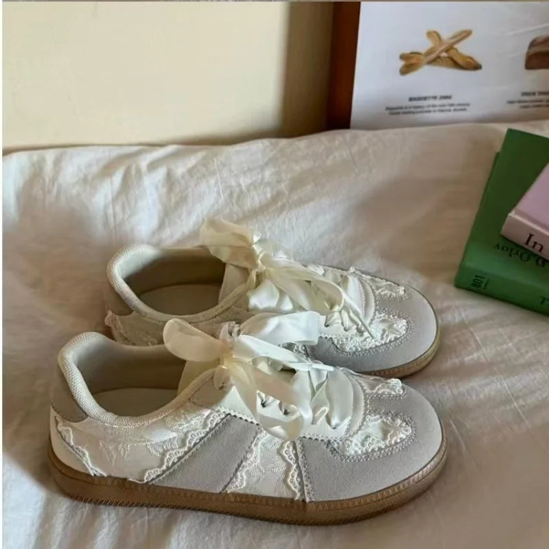A pair of low-top lace-up sneakers in off-white and light gray with floral lace appliques and wide satin ribbon laces, resting on a white fabric surface.