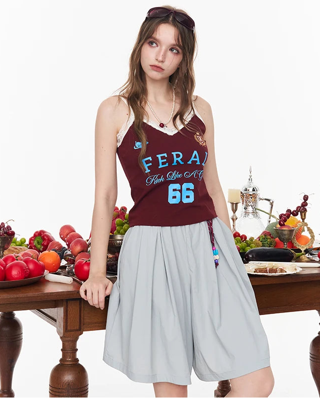 A woman models a sleeveless maroon tank top with 'FERAI Paris LE MONT 66' printed in light blue and white, paired with a light gray pleated skirt, posed next to a wooden table filled with fruit and tableware.