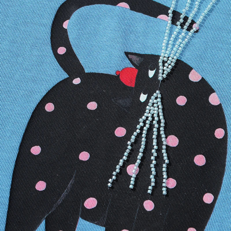 Close-up of a black cat graphic with pink polka dots on a light blue fabric, with a beaded necklace draped over the cat.