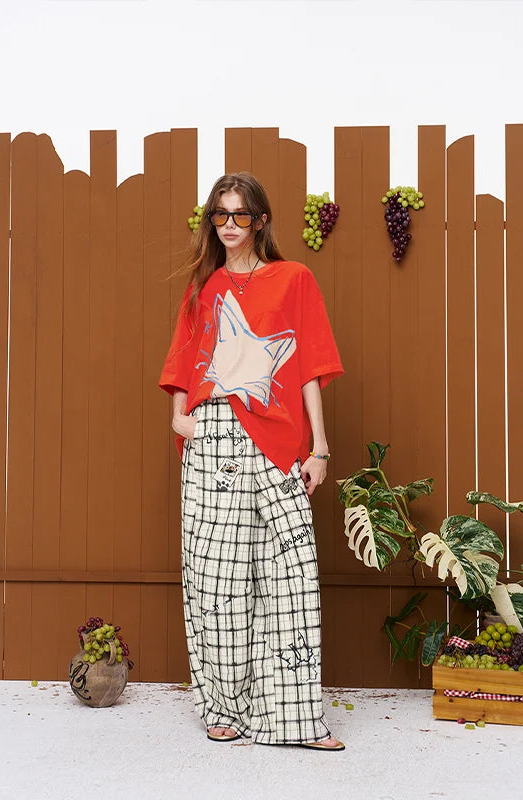 A woman stands in front of a brown wooden fence backdrop wearing a bright red oversized T-shirt with a large cream star graphic, wide-leg black-and-white plaid pants, and sunglasses. Decorative plants and grape props are placed around the set.