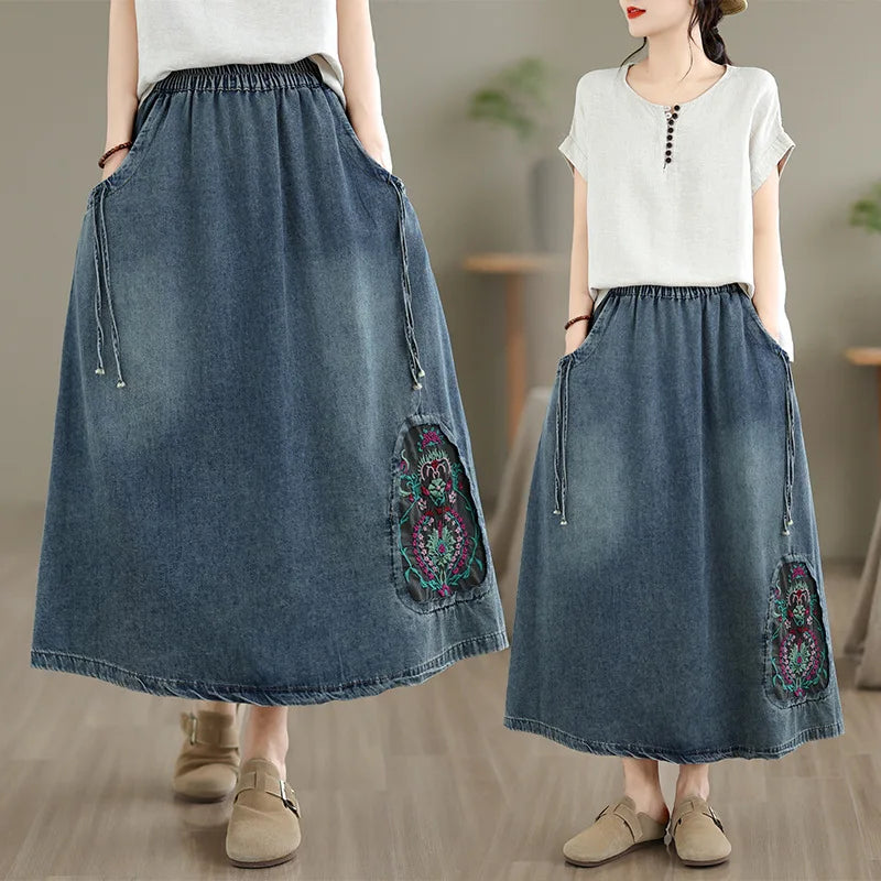 A model is shown wearing a long blue denim skirt with pockets and colorful embroidered details, paired with a white short-sleeve top. The image features a split view: a close-up on the left and a full-body shot on the right, set in a softly blurred indoor environment.