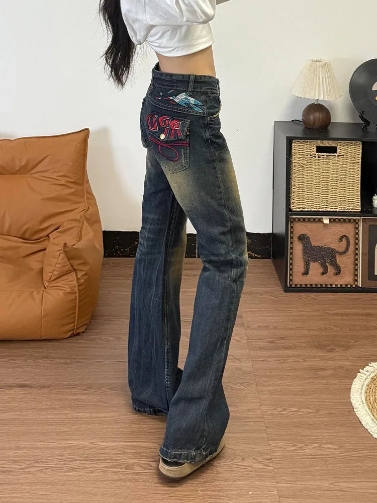 A person wearing dark blue flare jeans with colorful embroidered patches on the back pocket, standing in a room with a wood floor, a brown beanbag chair, and a small cabinet.