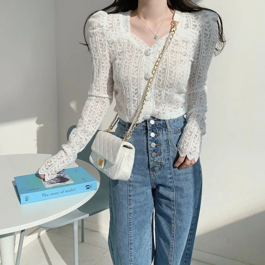 A woman wearing a white lace long-sleeve V-neck top and blue jeans, carrying a small white quilted shoulder bag with a gold chain strap, standing next to a white table with a blue book.
