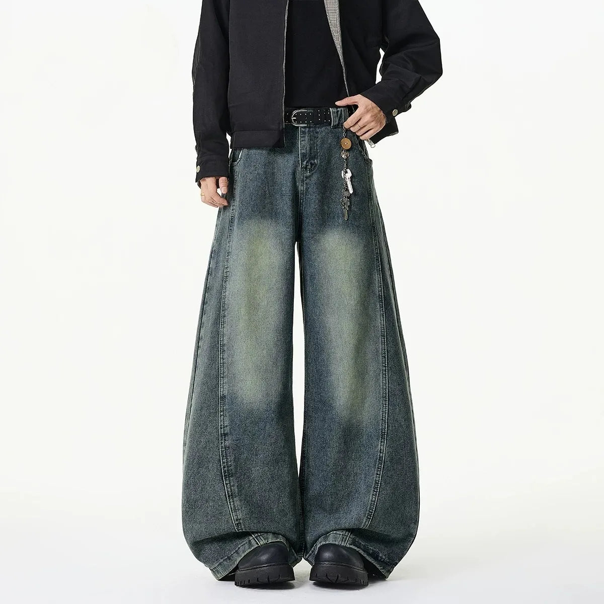 A person wearing very wide-leg faded blue denim jeans with a black jacket and black shoes, standing against a plain white studio background.
