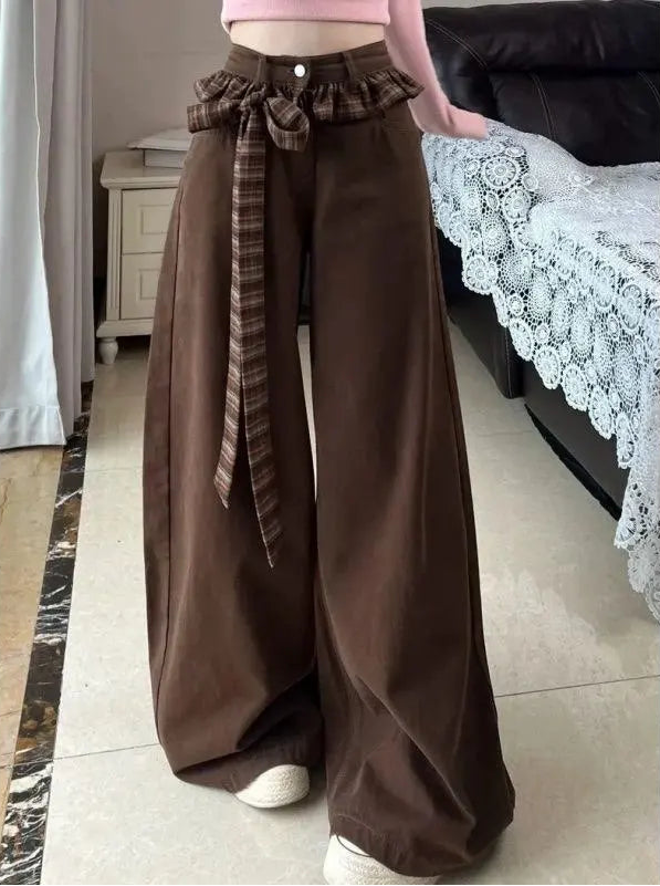 A person wearing wide-leg brown pants with a matching brown plaid tie belt and bow at the waist, paired with a pink cropped top and white sneakers in an indoor room setting.