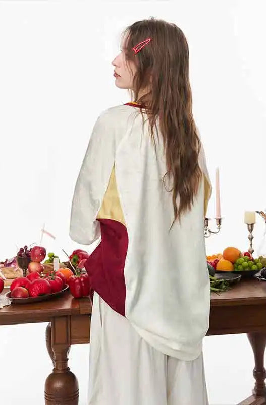 A woman with long brown hair is shown from the back wearing a loose, long-sleeve white dress with color-blocked panels in beige and deep red. She is standing in a studio setting next to a wooden table decorated with fresh fruit and candles.