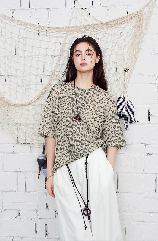 A woman stands against a white brick wall wearing a beige and black leopard-print short-sleeve top with white wide-leg pants. A fishing net hangs behind her, along with a striped fish decoration.