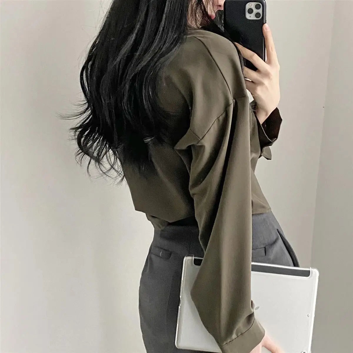 Person wearing an olive green shirt and gray pants, holding a phone and a white bag.