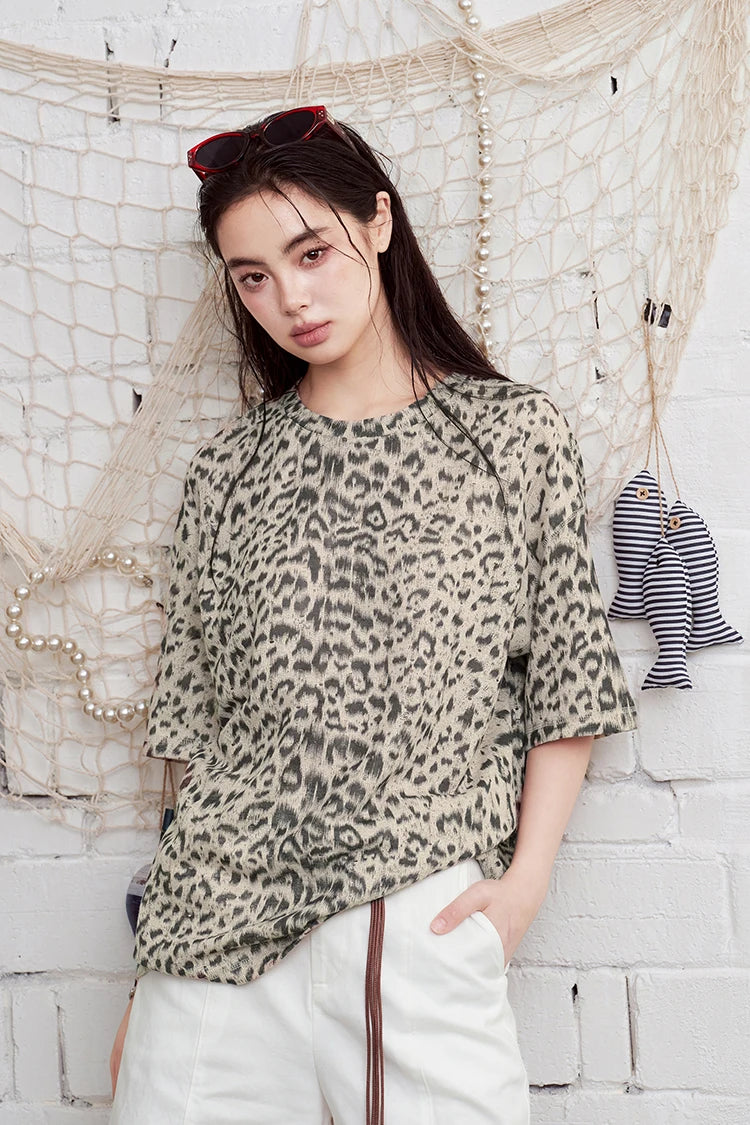 A woman wearing a beige and black leopard print short-sleeve top and white pants, standing against a white brick wall with a fishing net decoration.