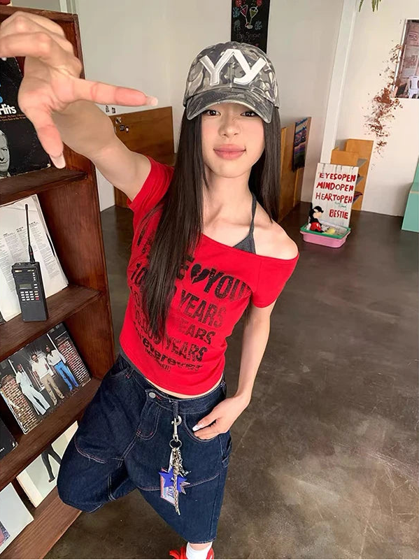 A woman posing indoors wearing a red off-the-shoulder graphic t-shirt, dark blue denim shorts, a camouflage baseball cap, and red sneakers. She is making a hand gesture in front of a wooden bookshelf.