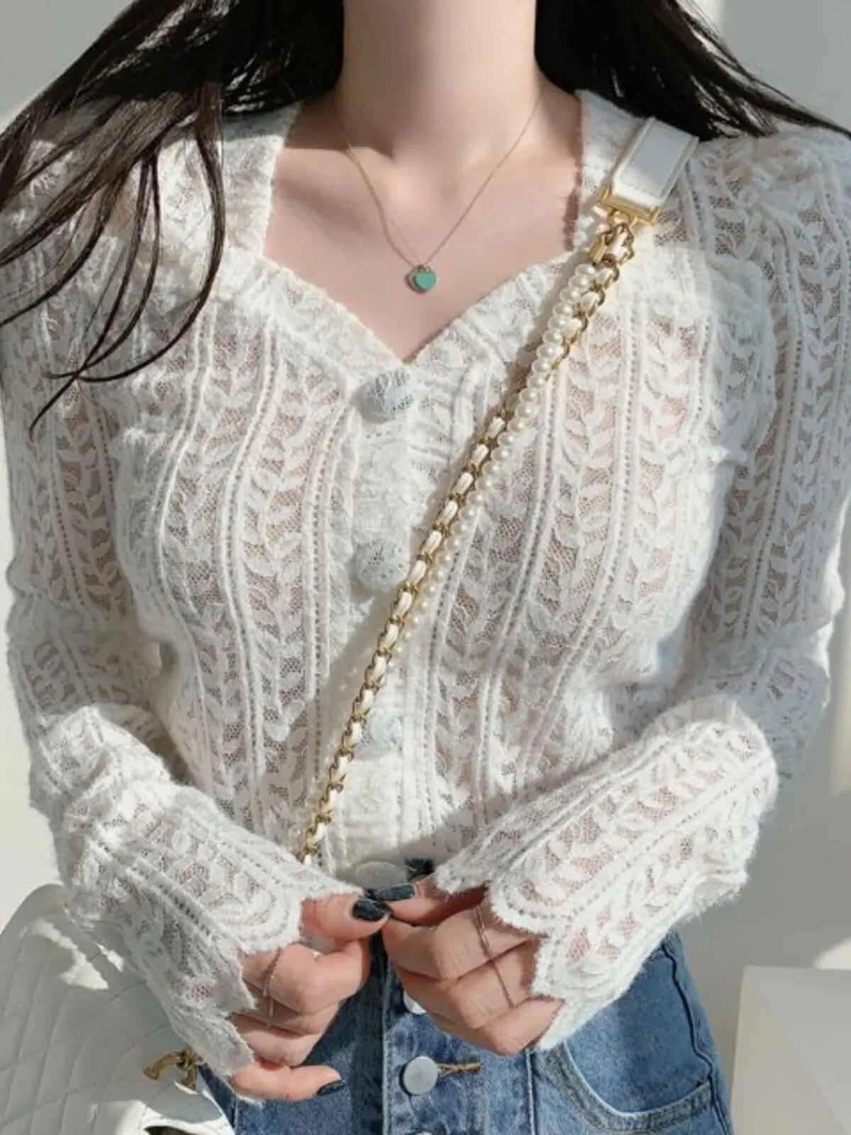 A close-up of a person wearing a white cable-knit V-neck cardigan with button details, paired with blue jeans and a gold chain strap bag.