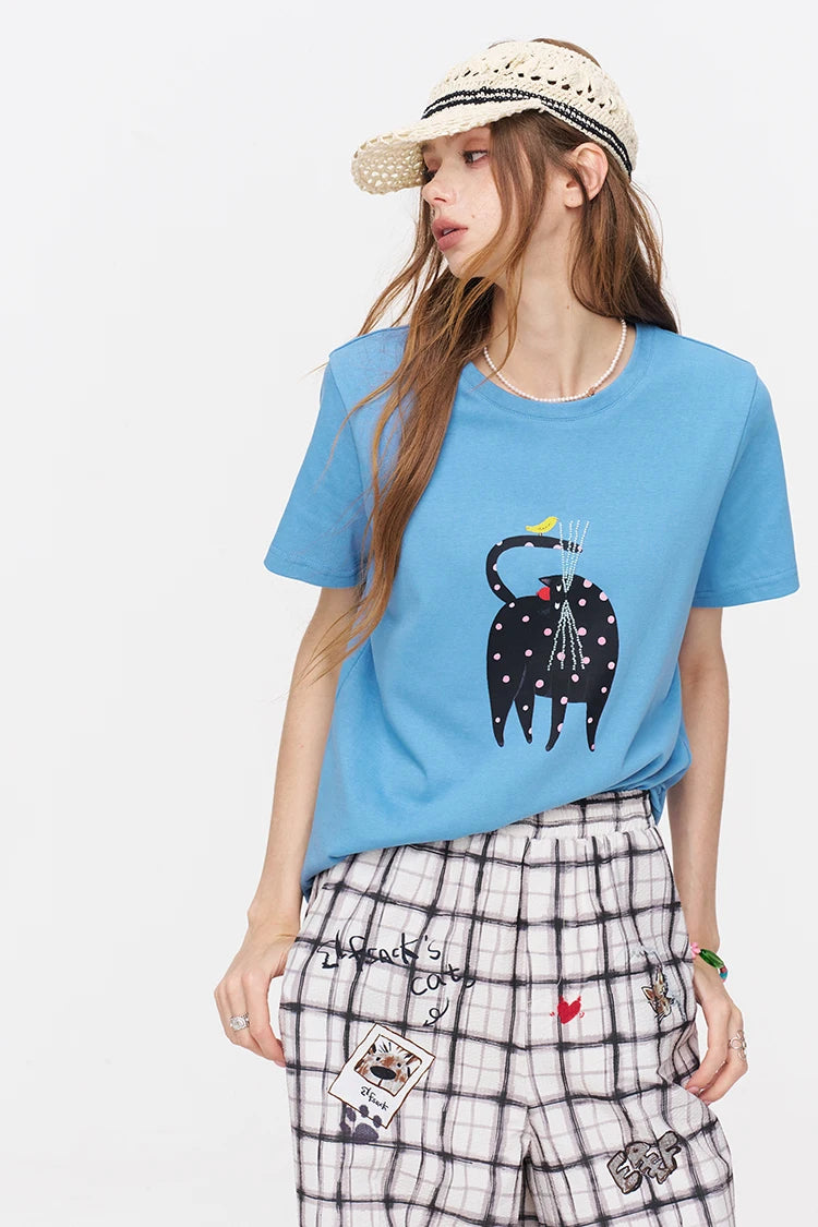 A woman modeling a light blue short-sleeve t-shirt with a black cat graphic, paired with black and white plaid shorts and a beige straw hat, against a plain white background.