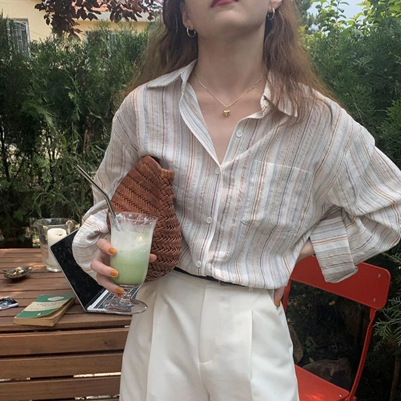 Woman holding a drink outdoors, wearing a striped shirt and white pants.