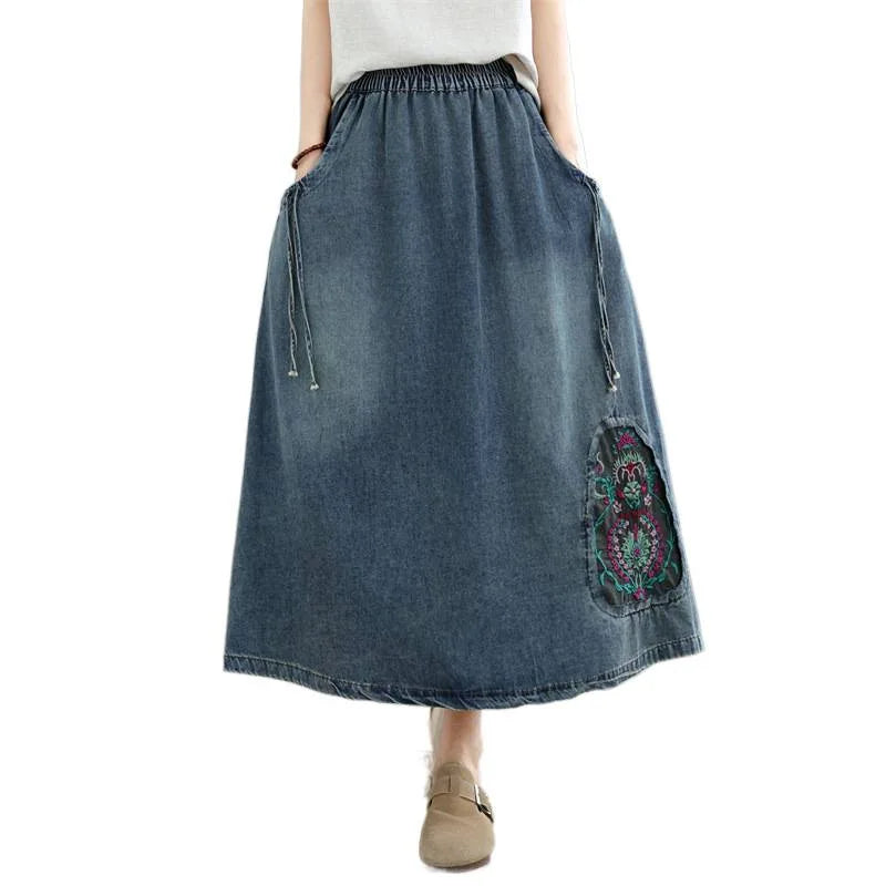 A model wearing a mid-length blue denim maxi skirt with a drawstring waist, side pockets, and a colorful embroidered patch on the lower right side, paired with a white top and beige loafers against a plain white background.