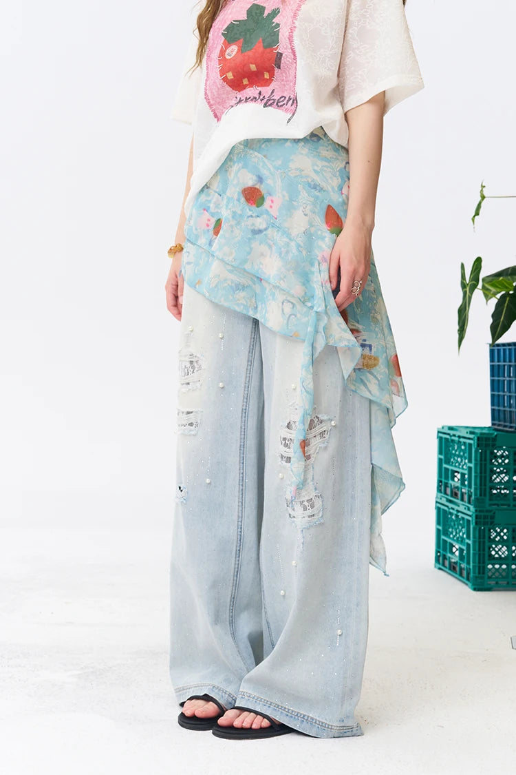 A model wearing a light blue distressed denim wide-leg pant with a layered, asymmetrical light blue floral shawl draped over the waist, styled with a strawberry-print t-shirt and black sandals.