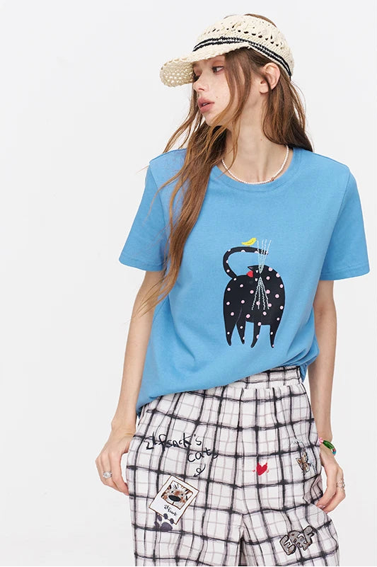 A woman modeling a light blue crewneck t-shirt with a black and white spotted elephant graphic on the front, paired with black and white plaid shorts, a woven straw hat, and a gold chain necklace, set against a plain white background.