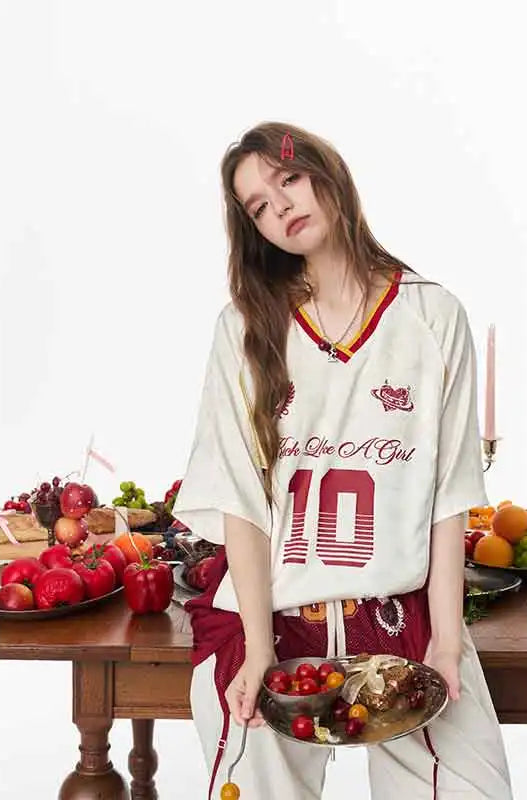 A woman models a cream-colored athletic jersey with red and yellow trim, featuring the text 'Just Like A Girl' and the number '10' on the front. She is holding a tray with food props in front of a table laden with fresh fruits, set against a clean white background.