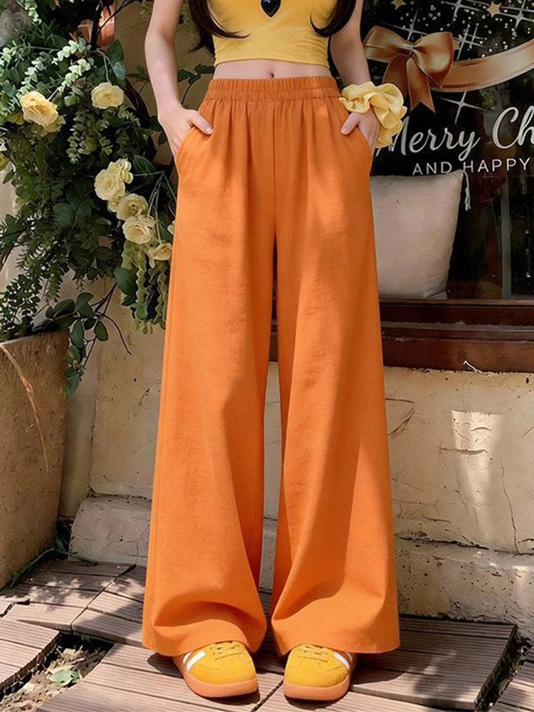 A person wearing bright orange wide-leg pants with a matching yellow cropped top and yellow shoes, standing outdoors near a stone wall and potted flowers.