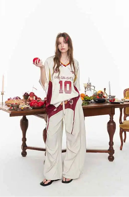 A woman stands in a studio setting wearing a cream and burgundy basketball-style lounge set with wide-leg pants and a matching top featuring the number 10. She holds a red apple beside a wooden table laden with fruit and candles against a plain white background.