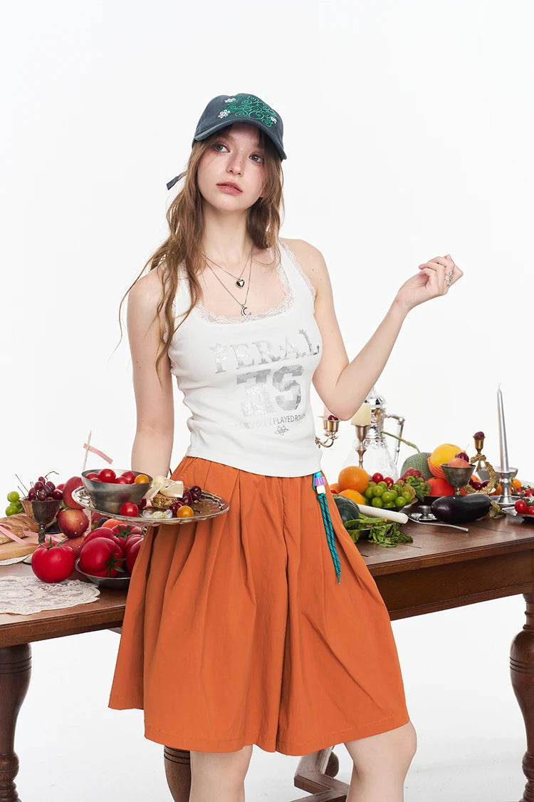 A woman modeling a white graphic tank top and an orange pleated skirt, standing behind a wooden table laden with fresh fruits and vegetables, wearing a green cap against a white studio background.