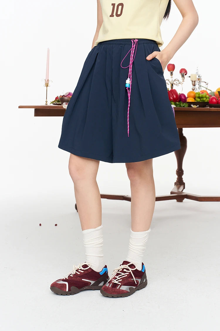 A person wearing a navy blue pleated mini shorts with a pink drawstring, styled with a cream t-shirt, white socks, and burgundy sneakers, standing in front of a white studio background with a wooden table and fruit.