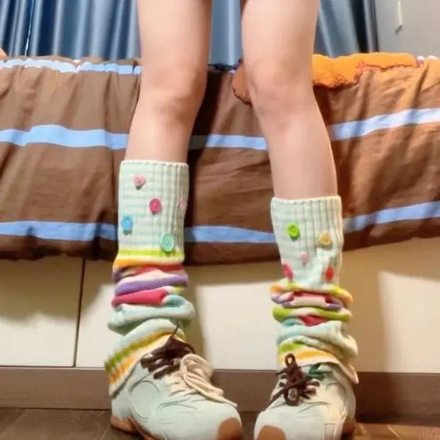 A person sitting on a bed wearing pastel multicolor stackable socks with colorful buttons and ribbed cuffs, paired with off-white sneakers.