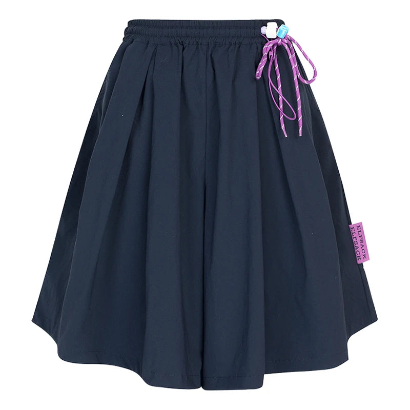 A dark navy blue pleated shorts with an elastic waistband, featuring a purple drawstring tied with a small white and blue flower accent on the right side and a purple brand tag near the hem.