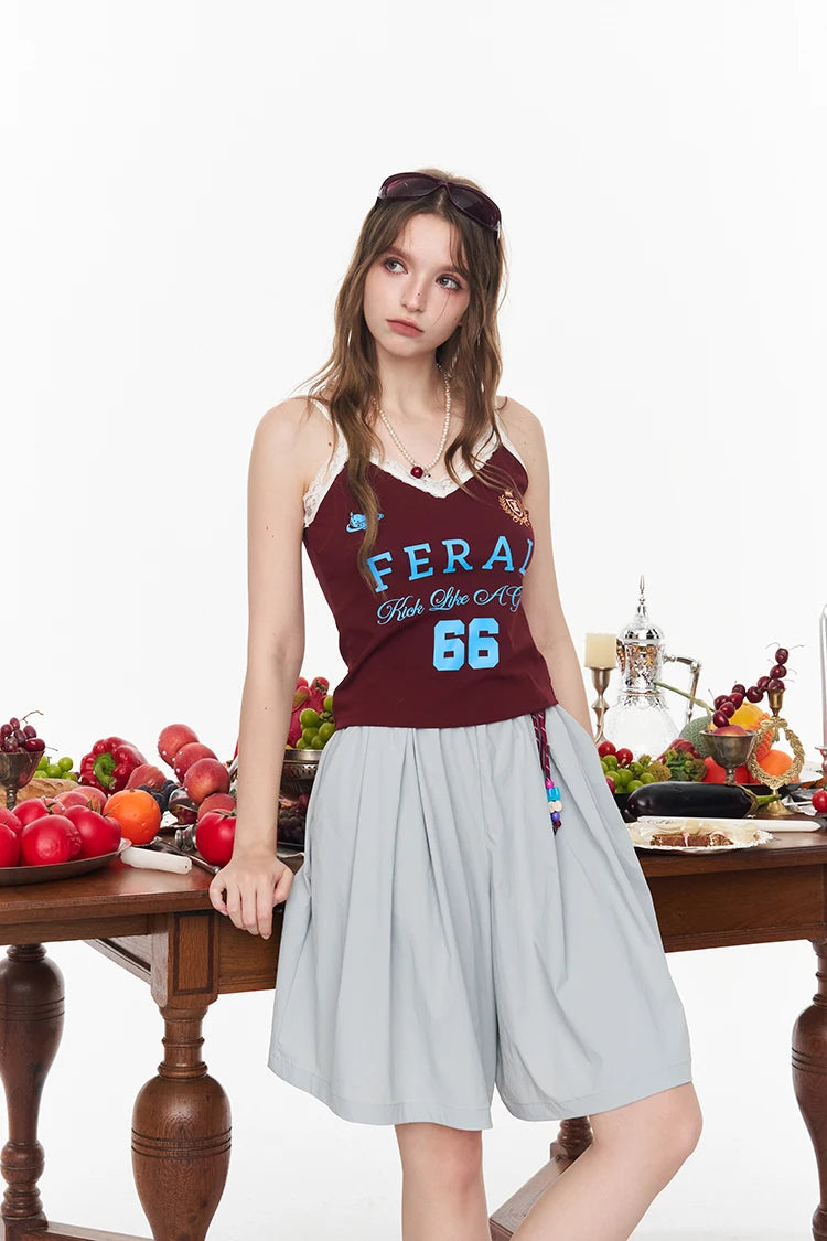 A woman models a sleeveless maroon tank top with 'FERA Fun Life Co 16' printed in light blue and white, paired with a light gray pleated shorts, posed behind a wooden table decorated with fresh fruit on a white studio background.