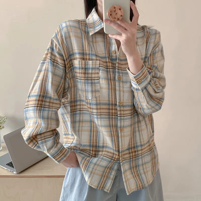 Person wearing a plaid shirt taking a mirror selfie in a room with a laptop on a desk.