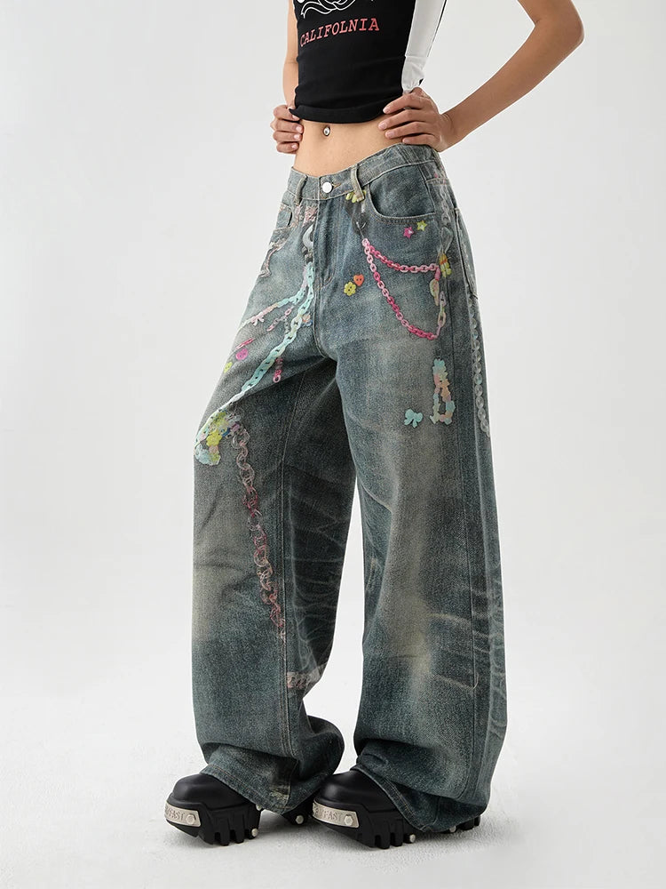 A model wearing a pair of wide-leg, heavily distressed denim jeans with colorful chain and bead embellishments, styled with a black crop top and chunky black platform sneakers against a light gray studio background.